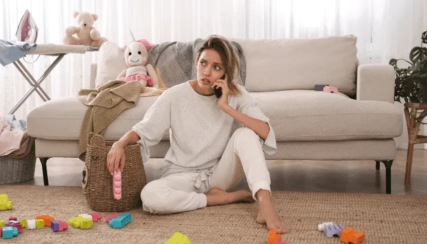 Stressed woman on the phone, sitting on the floor of a messy living room with toys, laundry, and an ironing board.