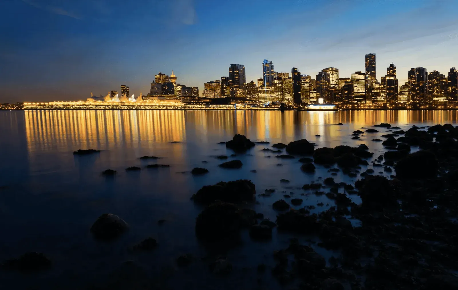 City skyline with warm lights reflecting on calm water at dusk, dark rocks in the foreground.