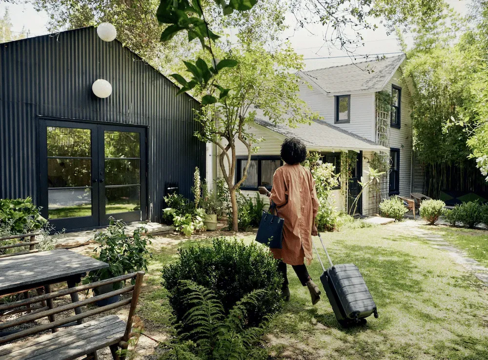 Person with curly hair and luggage walks through a lush green yard towards two houses, one dark modern, one white traditional.