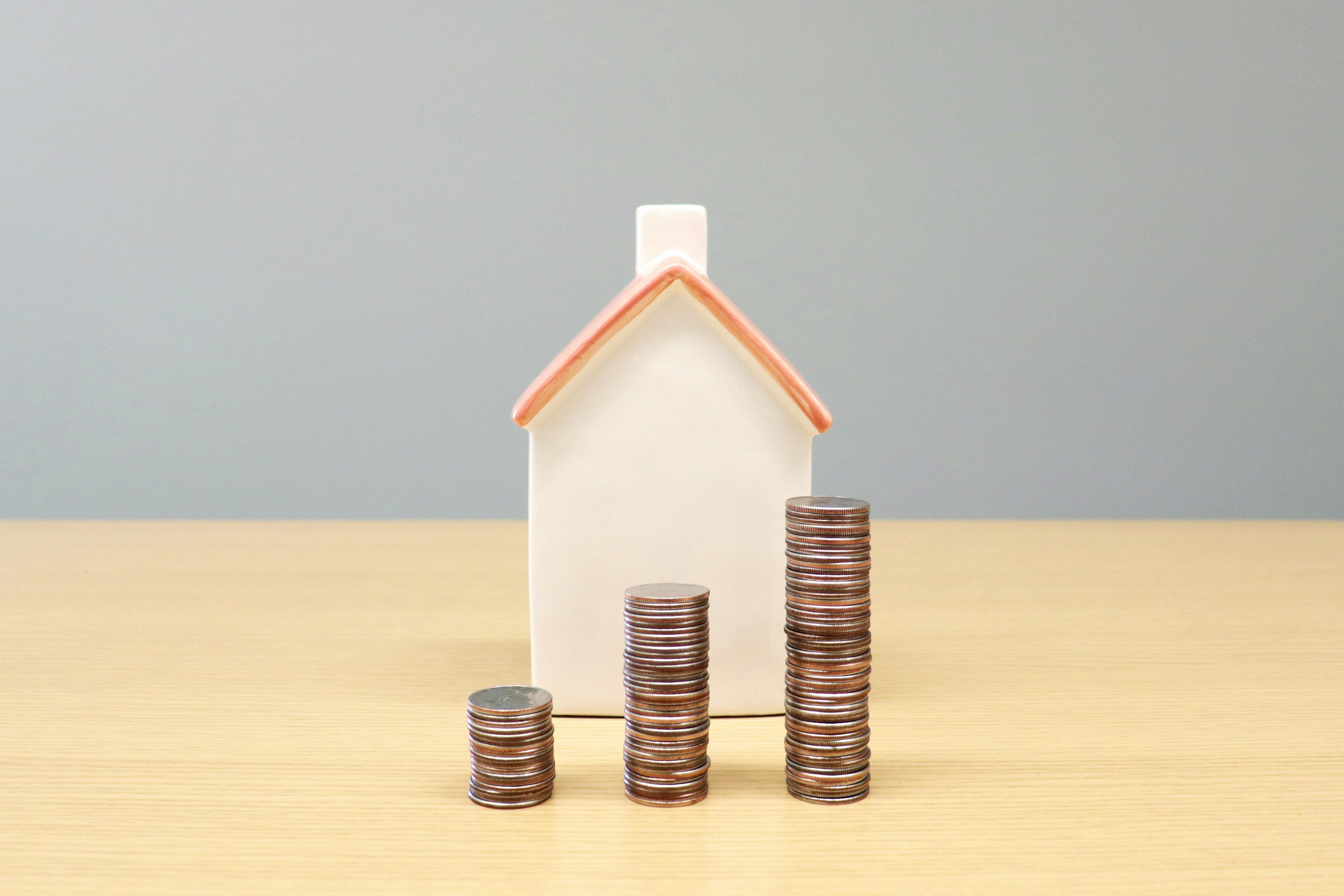 Three stacks of coins, increasing in height, in front of a small white house model with a red roof on a wooden table.
