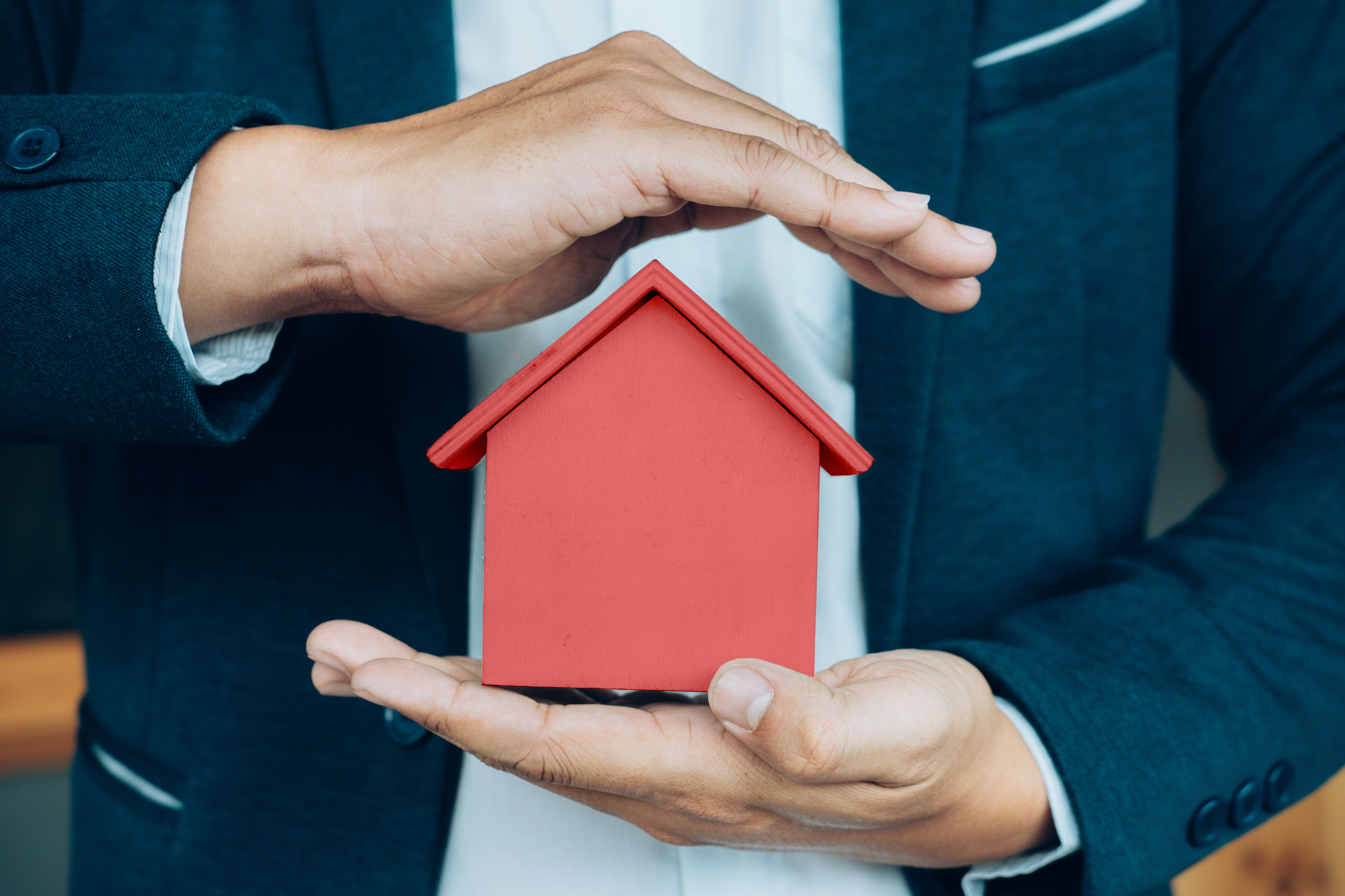 Hands in a dark suit hold a red house, with one hand protectively covering it from above.