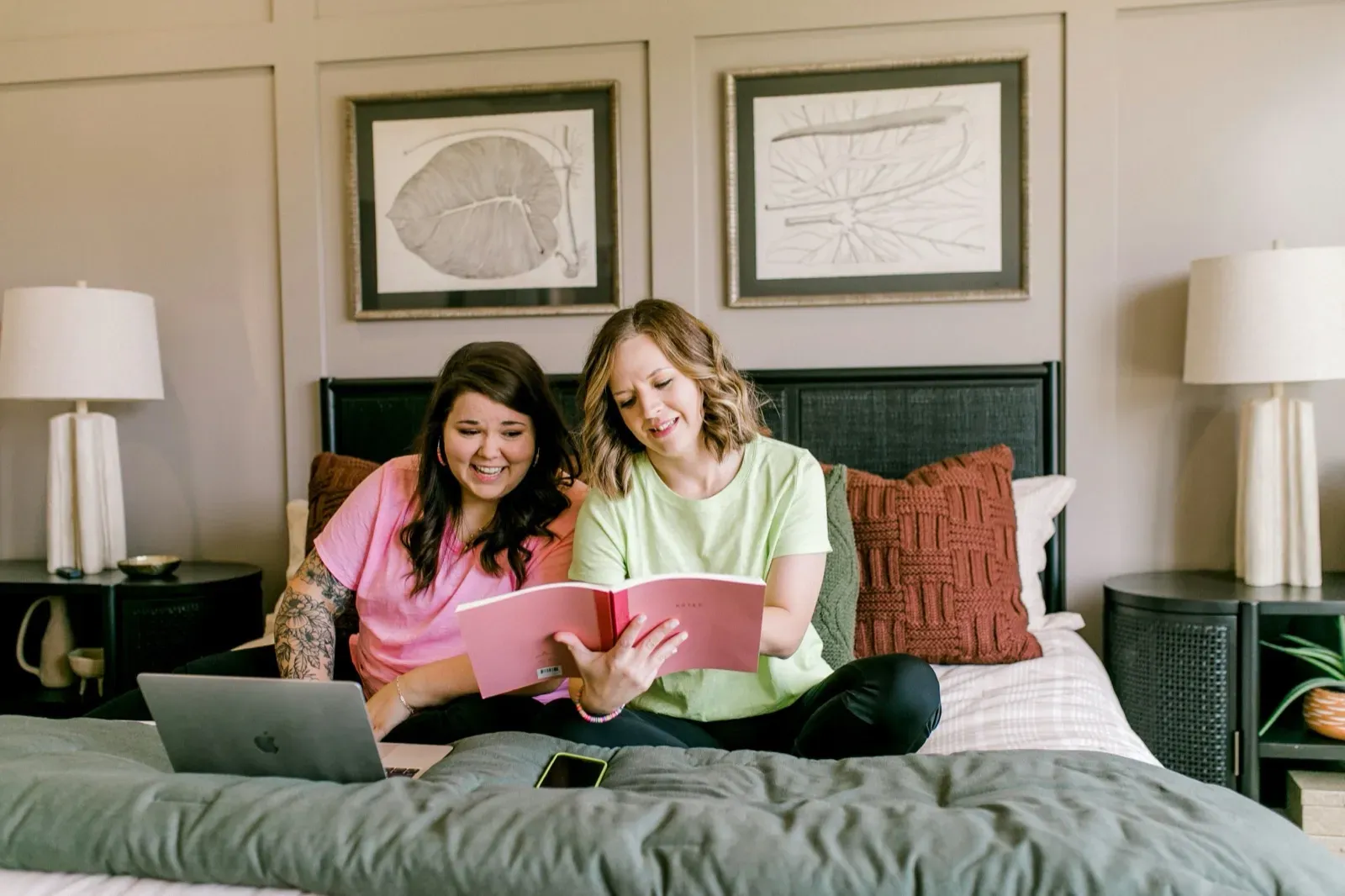Two women smile while relaxing on a bed; one uses a laptop, the other reads a pink notebook.