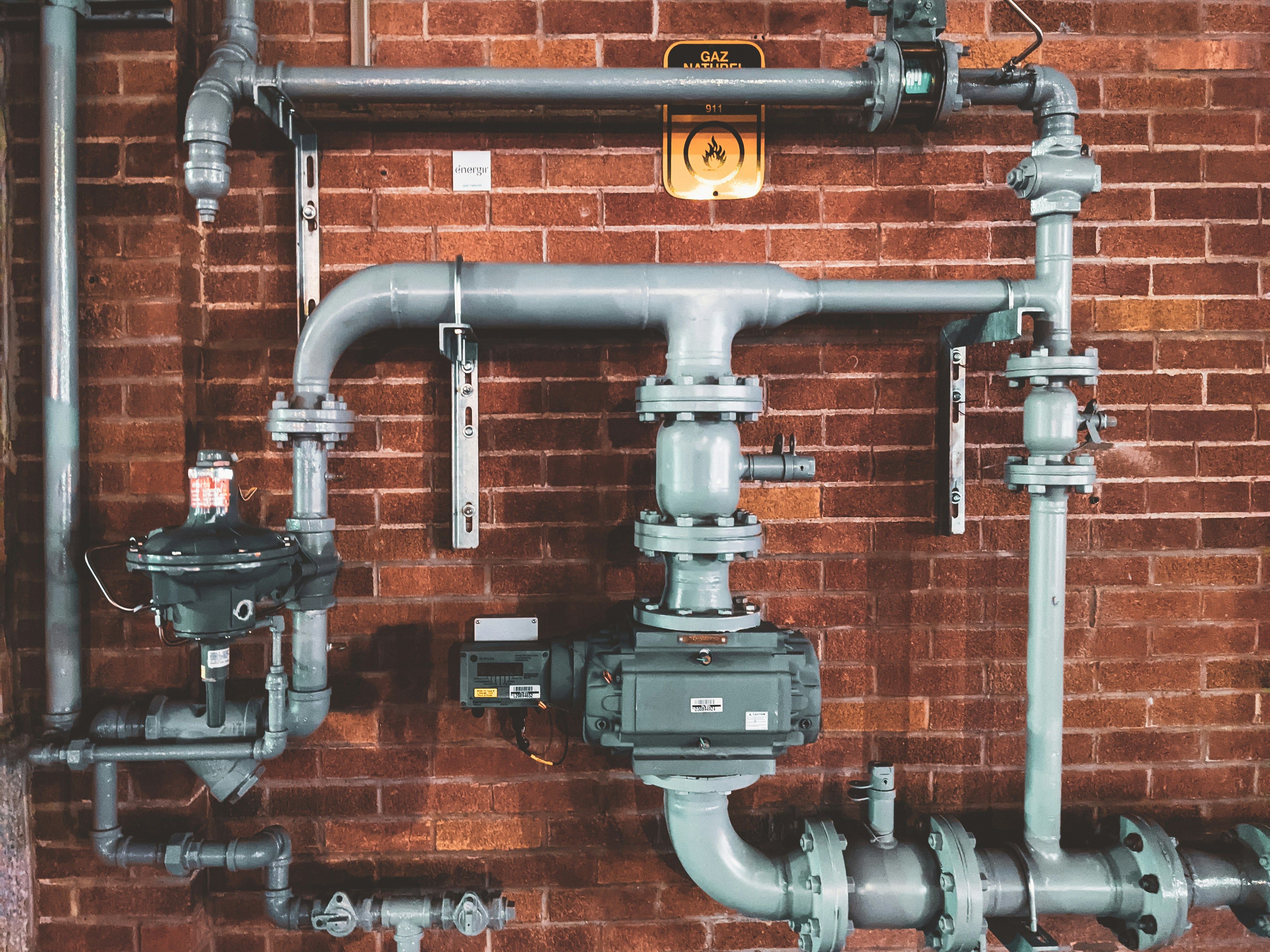 Complex gray industrial gas pipes, valves, and meters on a red brick wall with a "Gaz Naturel" sign.