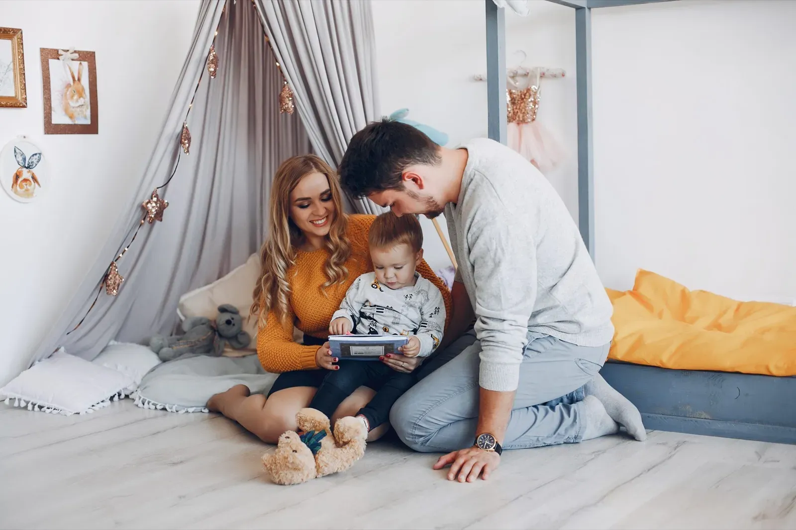 Happy family with toddler looking at a book on the floor in a bright, cozy bedroom.