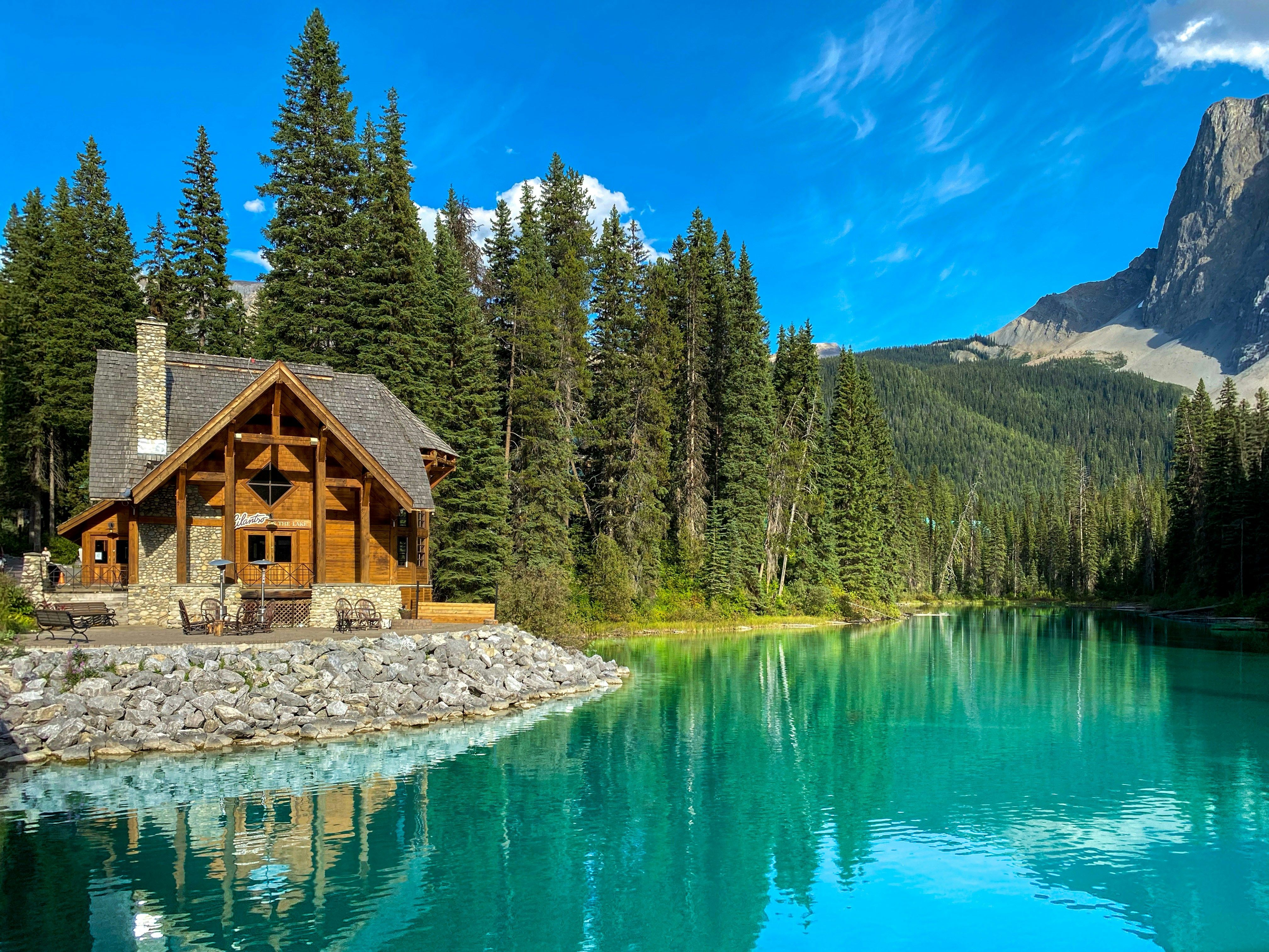 Log lodge by a vibrant turquoise lake, surrounded by tall evergreens, mountains, and a clear blue sky.