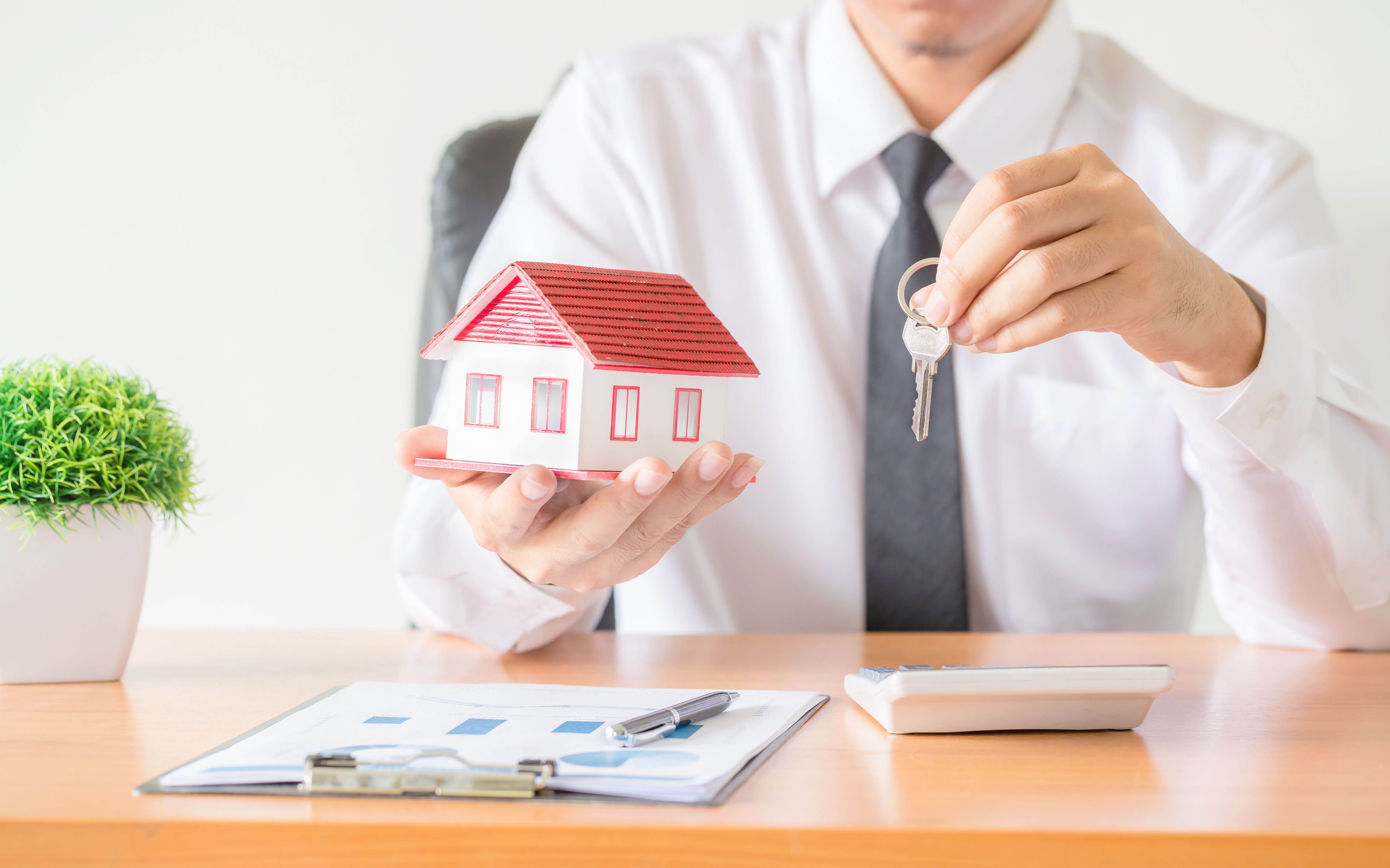Man in a white shirt and tie holds a miniature house model and house keys, sitting at a desk with documents.
