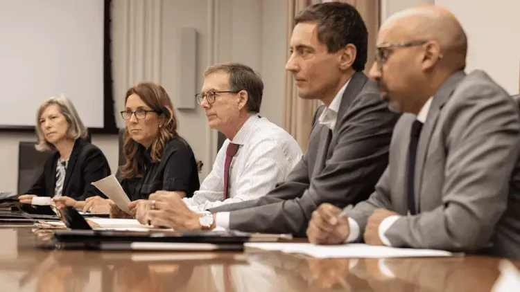 Five professionals (3 men, 2 women) sit attentively at a conference table with documents.