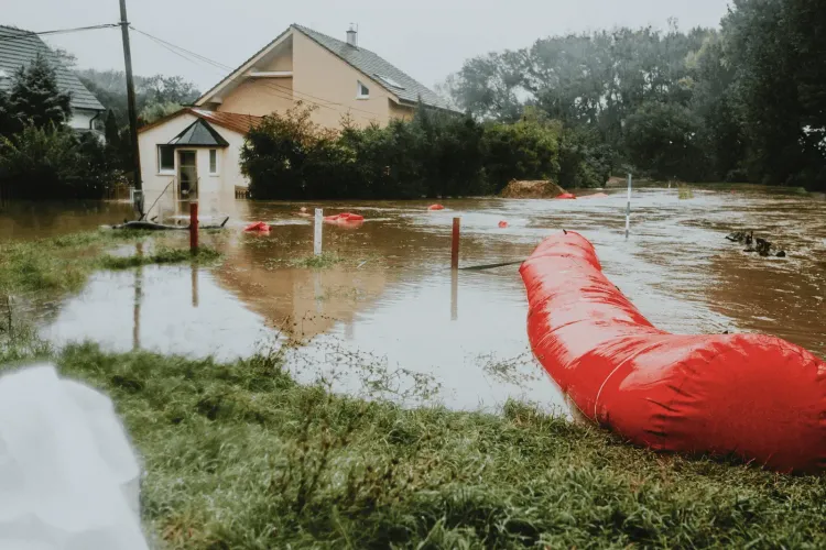 Houses partially submerged in muddy floodwaters with a large red flood barrier in the foreground.