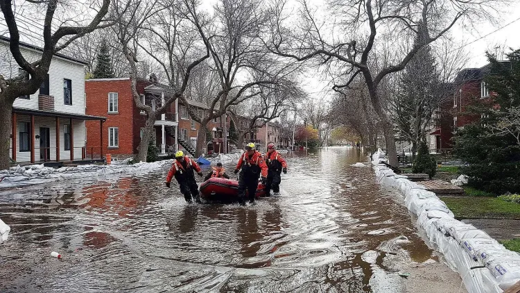 Rescue workers push a boat with a person through a flooded street lined with sandbags and houses.