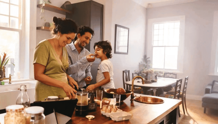 Happy family cooking in a bright kitchen. Dad feeds their child a snack while mom preps ingredients.