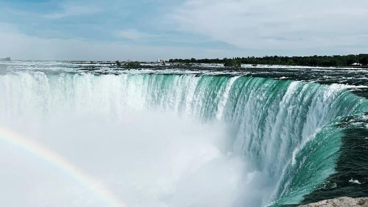 Powerful Niagara Falls, showing its curved turquoise water cascading into mist, with a faint rainbow below.