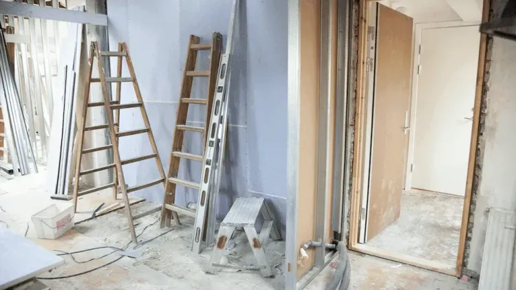 Room under renovation with two wooden ladders, exposed wall framing, and construction materials on a dusty floor.
