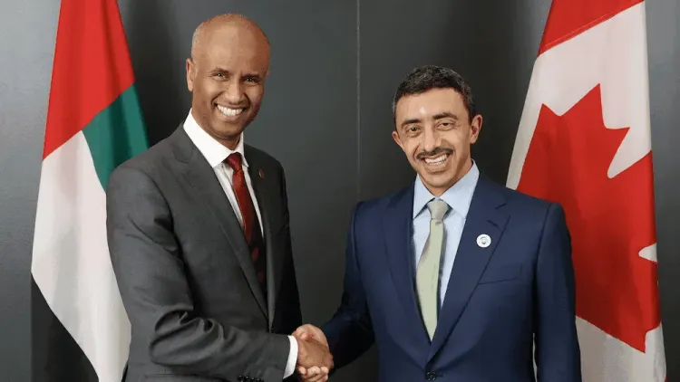 Two men in suits smiling and shaking hands, with UAE and Canadian flags behind them.