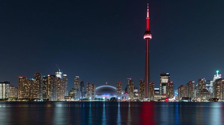 Toronto skyline at night with the CN Tower glowing red, Rogers Centre dome, and city lights reflecting on water.