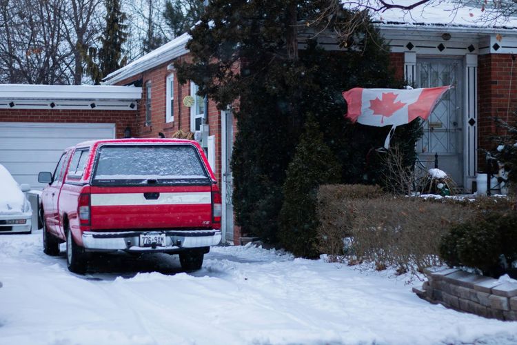 Red pickup truck in a snowy driveway with a Canadian flag on a brick house during a light snowfall.