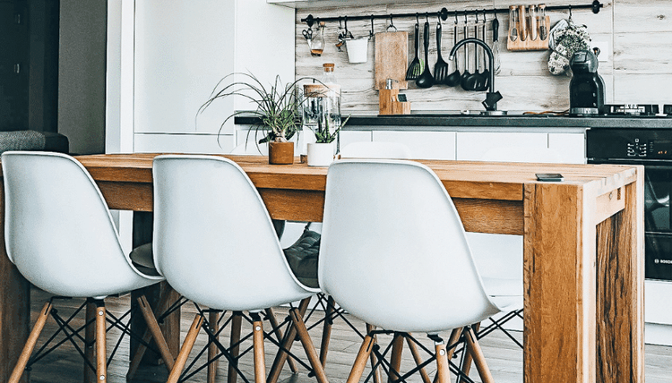 Modern kitchen with a long wooden dining table, three white chairs, plants, and kitchen tools hanging on the wall.