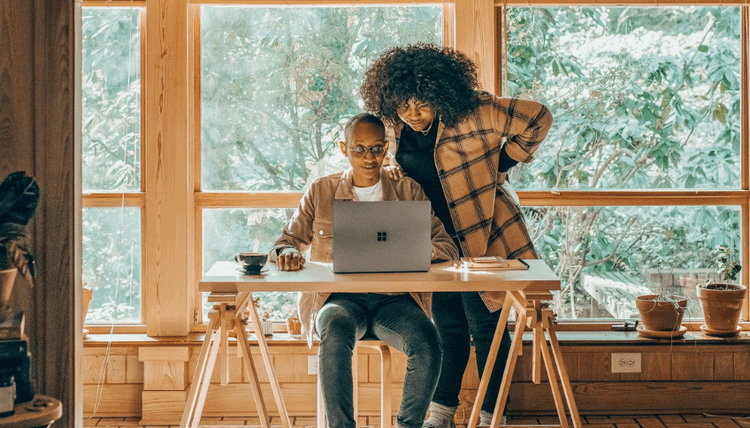Two people collaborate on a laptop at a desk by large windows with trees outside.