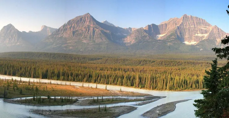 Panoramic mountain landscape: snow-capped peaks, dense forest, and a broad, winding river.