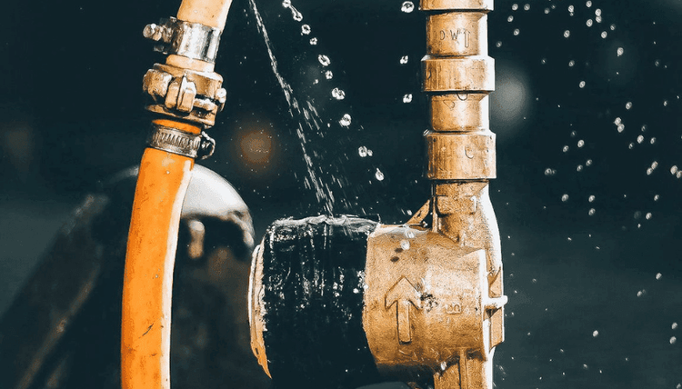 Water sprays from a leaky connection between an orange hose and a brass pipe, partly covered with black tape.