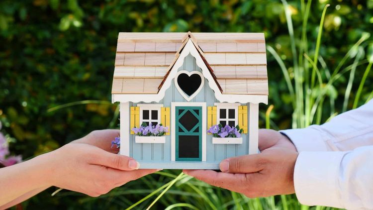 Two hands hold a blue miniature house with a heart-shaped window, yellow shutters, and purple flowers.