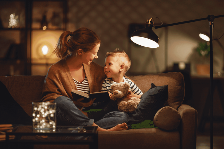 Smiling mother reads a book to her young child, who holds a teddy bear, on a couch.