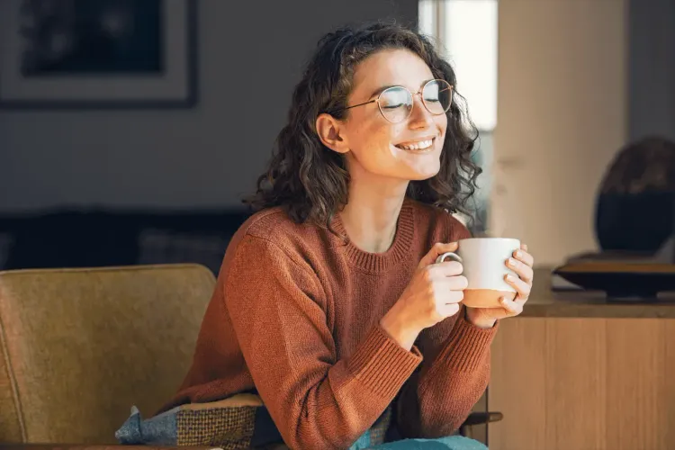 Smiling woman in glasses with curly hair holds a steaming mug, eyes closed in contentment.