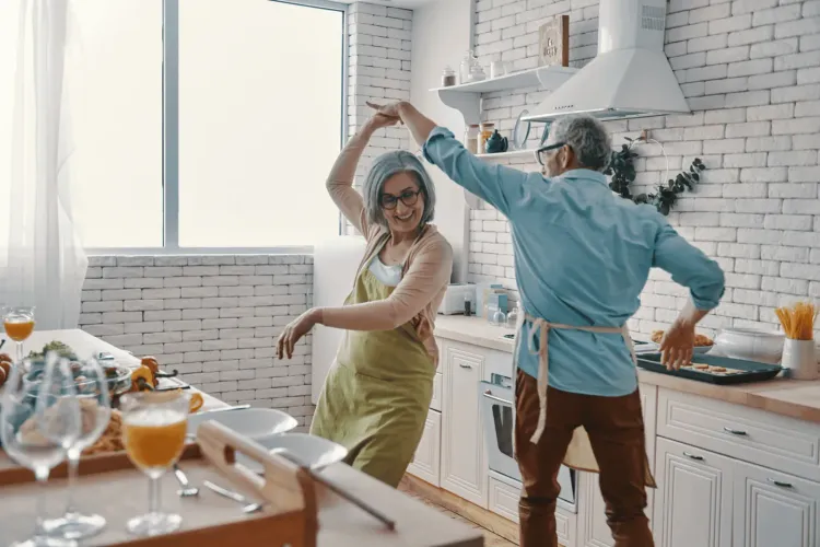 Cheerful senior couple in aprons dancing together in a bright, modern kitchen.