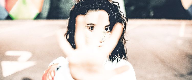 A woman with wet dark hair holds her hand up, palm out, largely obscuring her face, one eye looking intently at camera.