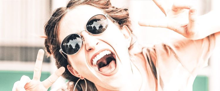 Excited woman in round sunglasses (trees reflected), mouth wide open, making peace sign & "okay" sign.
