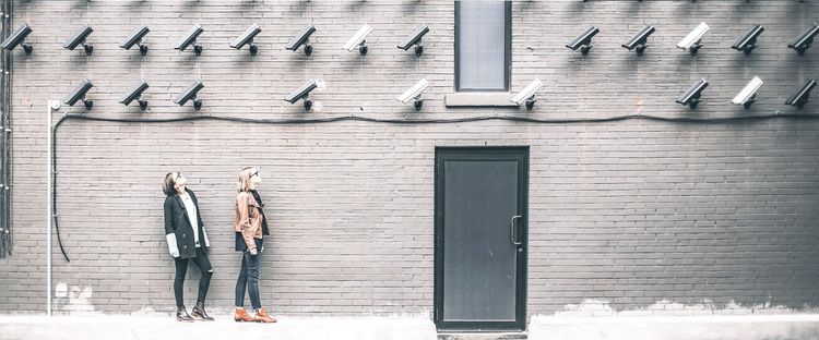 Two women look up at a brick wall covered with numerous surveillance cameras, alongside a door and window.