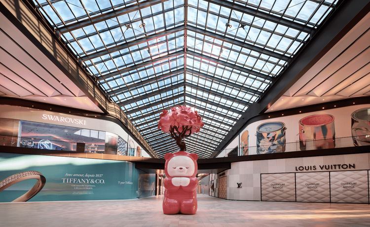 Large glossy pink bear sculpture with a pink tree on its head in a modern shopping mall with a glass roof.