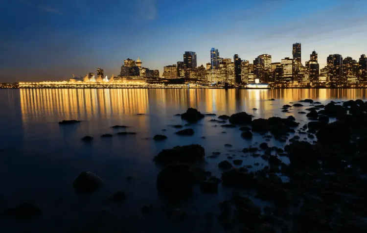 City skyline with warm lights reflecting on calm water at dusk, dark rocks in the foreground.