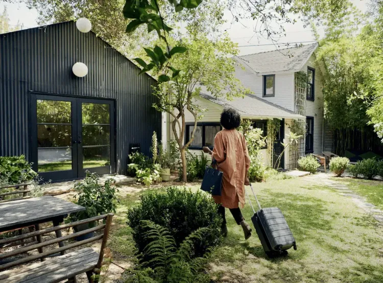Person with curly hair and luggage walks through a lush green yard towards two houses, one dark modern, one white traditional.