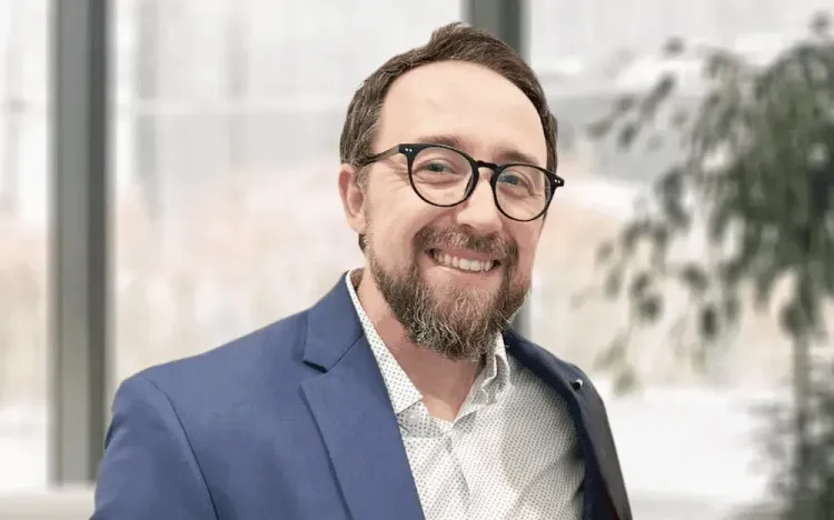 Smiling bearded man in glasses and a blue suit jacket, with a blurred office window background.