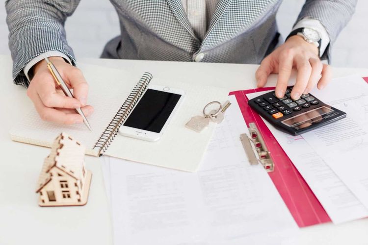 Person in a suit writes in a notebook and uses a calculator, next to a house model, keys, and phone on a desk.