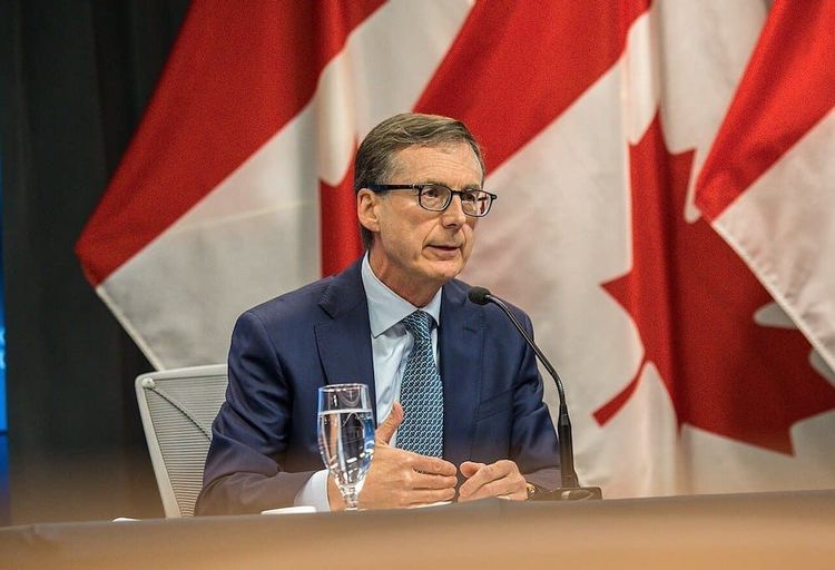 Man in blue suit and glasses speaks at a microphone, hand gesturing, with two Canadian flags in the background.