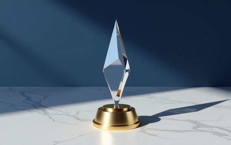 A sharp, clear crystal trophy on a gold base on white marble with a blue background and sunlight shadows.