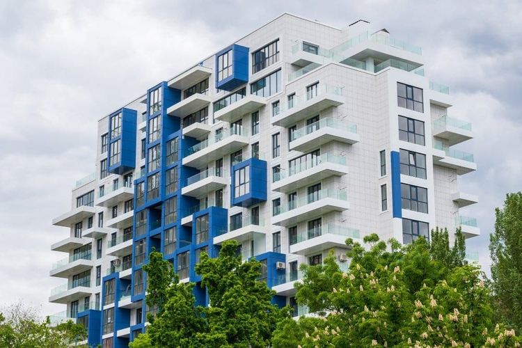 Modern white and blue apartment building with balconies and glass railings, with green trees in the foreground.