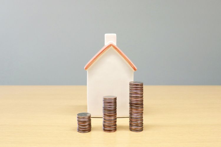 Three stacks of coins, increasing in height, in front of a small white house model with a red roof on a wooden table.