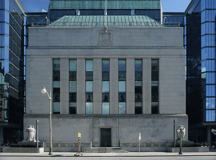 Grey stone neoclassical building with Royal coat of arms, flanked by blue glass towers under clear sky.