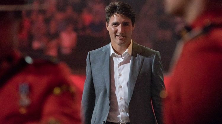Justin Trudeau smiling in a grey suit and white shirt against a blurred red background.