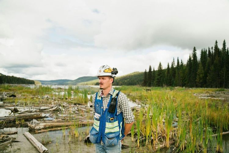Man in hard hat and work vest stands in shallow water with floating logs, tall grass, and a distant forest.