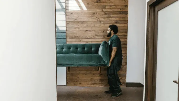 A man carrying a green velvet tufted sofa through a room with a wooden accent wall.