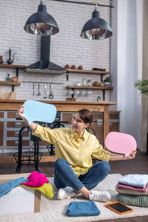 A happy woman sits on the floor holding up blank blue and pink speech bubbles, with clothes for donation.