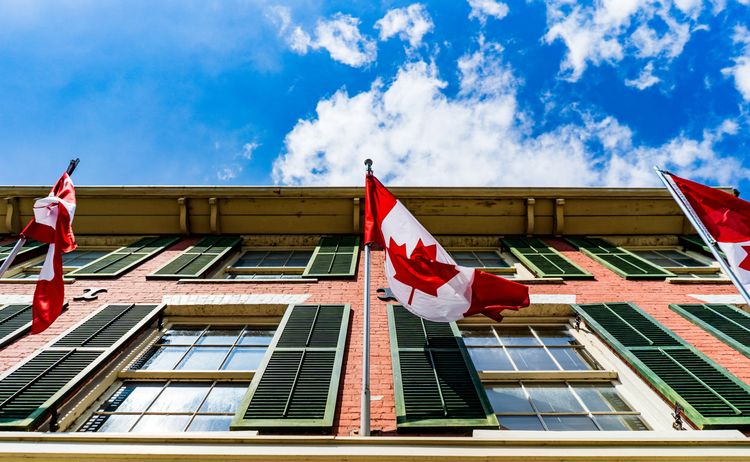 Three Canadian flags waving above a red brick building with green shutters against a bright blue sky.