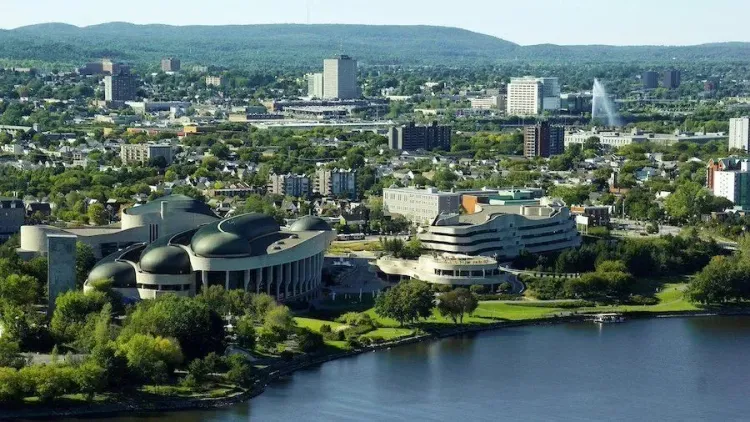 Aerial view of the Canadian Museum of History next to the Ottawa River, with Ottawa city and treed hills in the background.