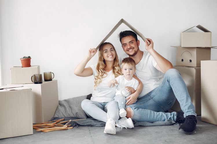 Smiling family with a child sitting on the floor surrounded by moving boxes, holding a cardboard house shape over their heads.
