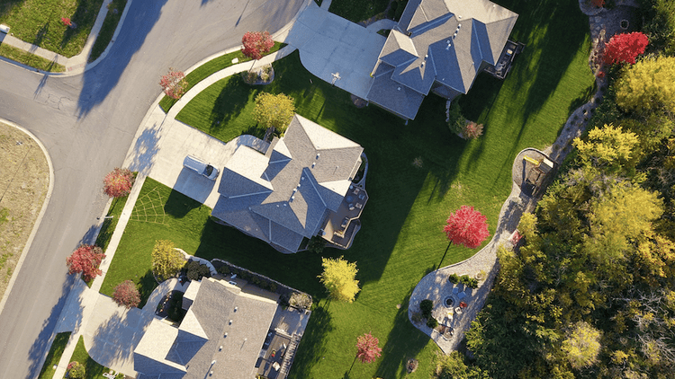 Overhead drone shot of a sunny suburban neighborhood with houses, green lawns, streets, and vibrant autumn trees.