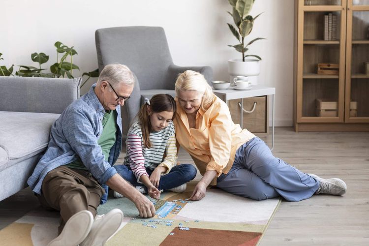 Grandparents and granddaughter happily doing a jigsaw puzzle together on a colorful rug in a bright living room.