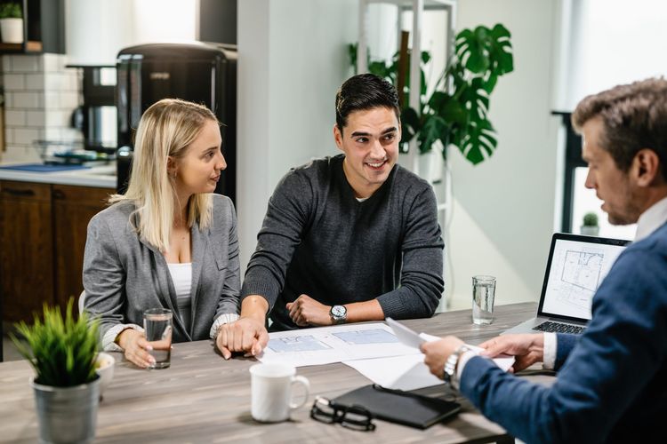 A couple holds hands, smiling, as they meet with a consultant discussing documents and a laptop with a floor plan.
